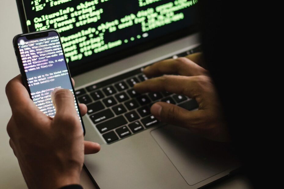 Free Unrecognizable hacker with smartphone typing on laptop at desk Stock Photo